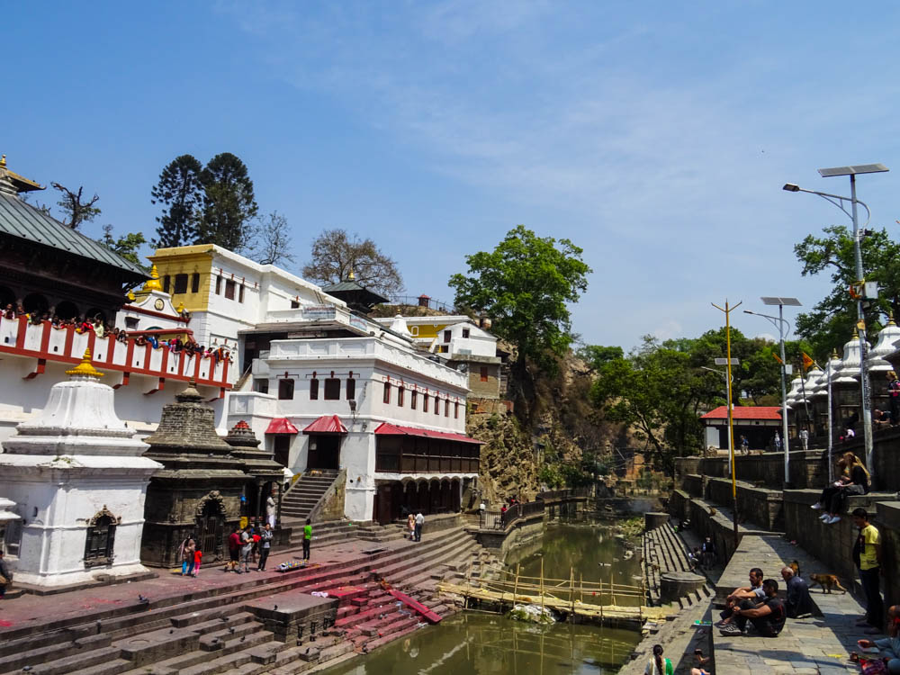 Pashupatinath mit Schreinen rechts des Flusses Bagmati. Auf der linken Seite des Flusses ist ein Tempel, davor ist ein Verbrennungsplatz zu sehen.