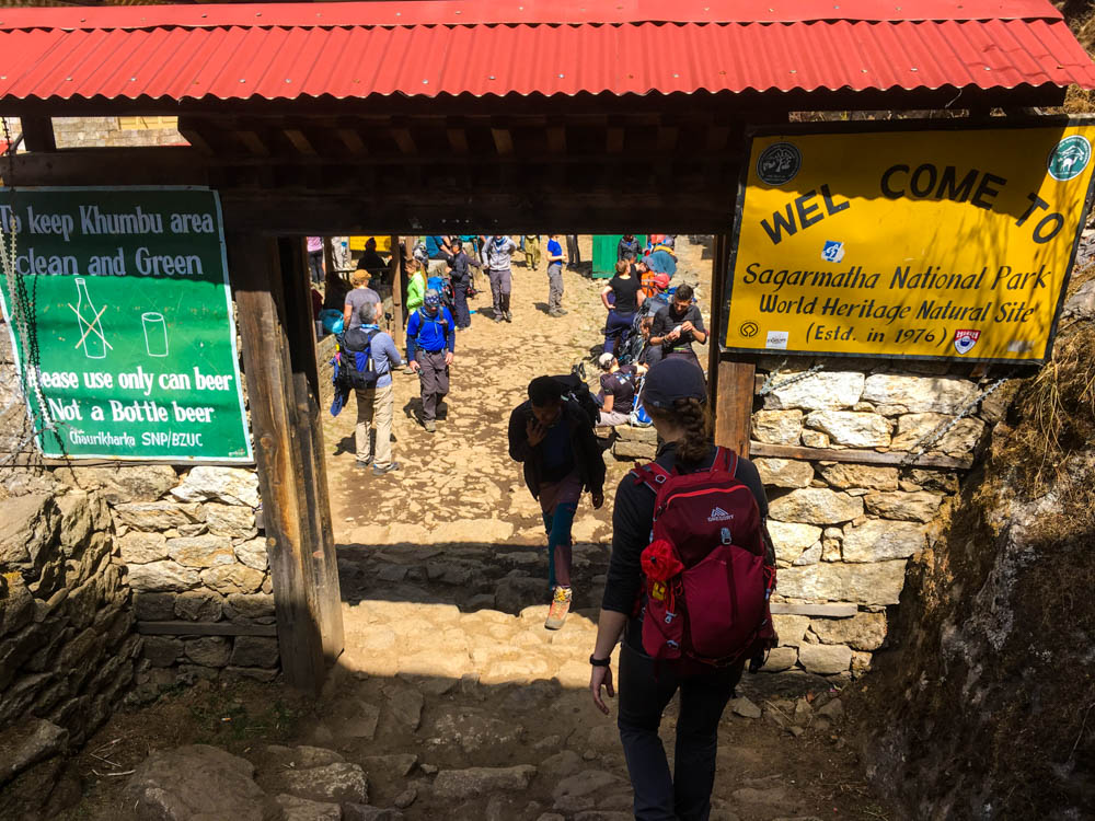 Melanie läuft durch das Eingangstor zum Sagarmatha Nationalpark. Rechts davon ist ein Schild das einen Wilkommen heißt und erklärt, dass der Park seit 1976 Weltnaturerbe ist. Rechts ist ein Schild, dass ab dieser Stelle Glasflaschen verboten sind und man Bitte Dosenbier statt Bier aus der Flasche trinken soll.