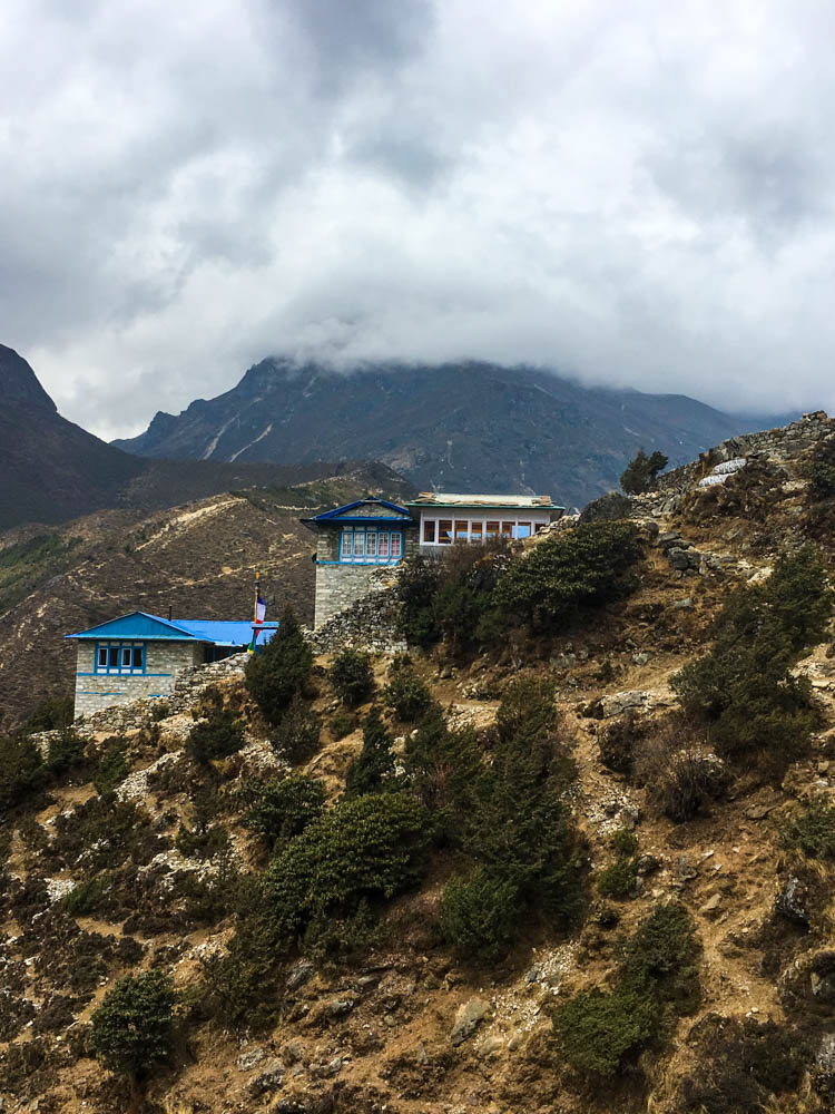 Klosteranlage Thame in Nepal. Es sind ein paar Gebäude zu sehen, ansonsten ein Bergrücken im Hintergrund. Die Gipfel sind mit Wolken behangen.