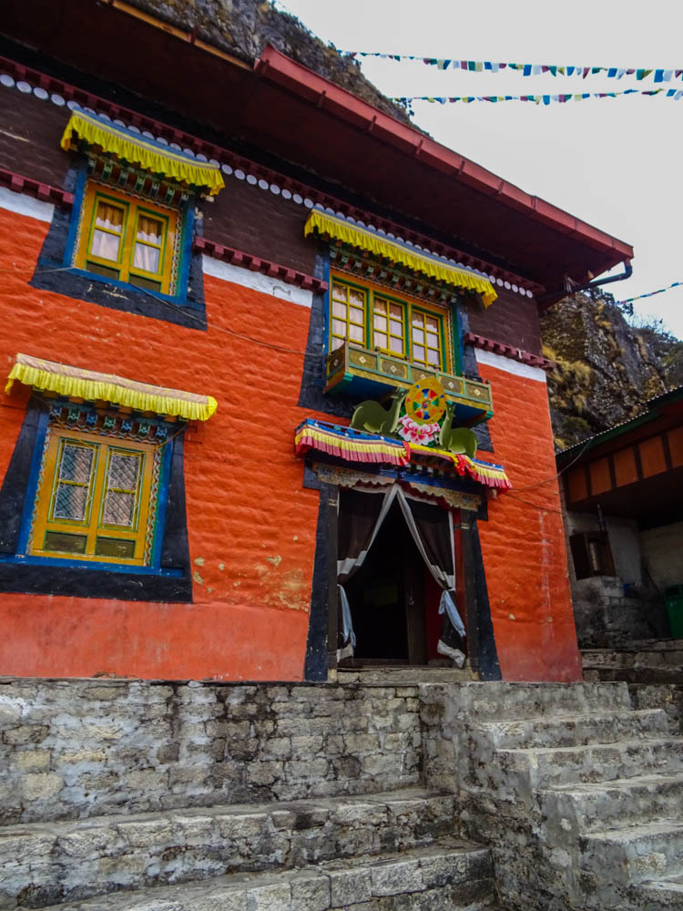 Bunte Front eines Klosters in Thame. Über den blau-gelben Fenstern sind gelbe Tücher angebracht. Die Hauswand ist rot-orange. Am Himmel sind bunte Gebetsfahnen zu sehen.
