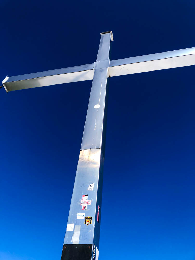 Das Kreuz wird von der Sonne in den Alpen angestrahlt, der Himmel drum herum ist kräftig blau.
