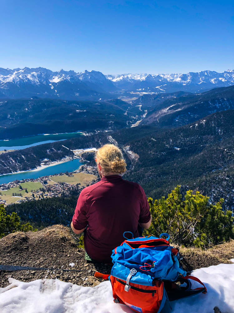 Julian macht eine Pause am Martinskopf und sitzt am Rande des Gipfels. Ausblick auf Walchensee und Bergpanorama. Der Himmel strahlt blau.