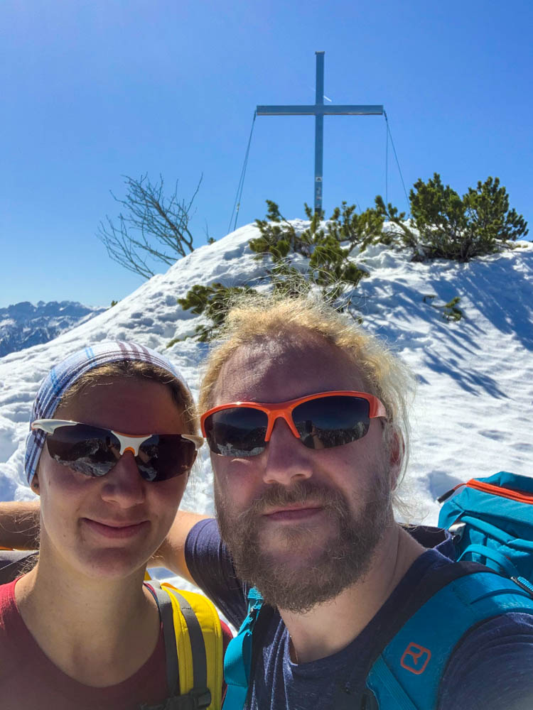 Selfie von Melanie und Julian am Martinskopf. Hinter ihnen ist das Gipfelkreuz zu sehen. Die Sonne in den Alpen lässt den Schnee rund herum strahlen, der Himmel ist blau.