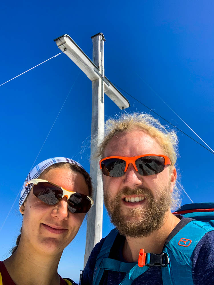 Selfie von Melanie und Julian mit Gipfelkreuz von Herzogstand. Der Himmel strahlt kräftig blau.