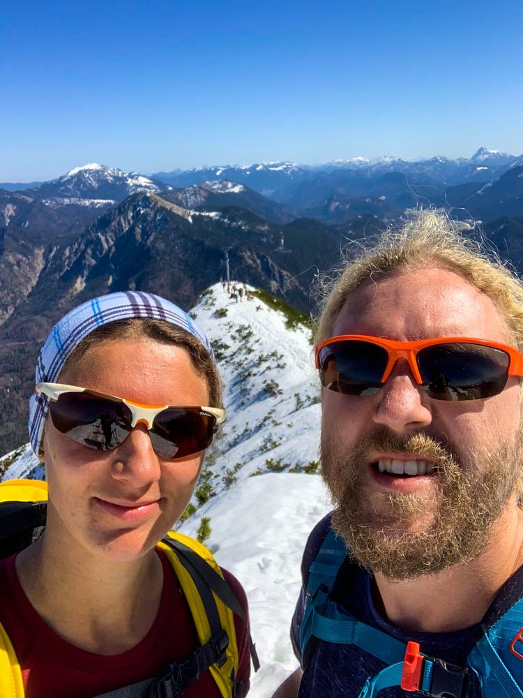 Selfie von Melanie und Julian. Zwischen ihnen etwas unterhalb ist der Gipfel des Herzogstand zu sehen, sowie die dahinterliegende Berglandschaft. Die Sonne in den Alpen verwöhnt die beiden und der Schnee ist strahlend weiß.