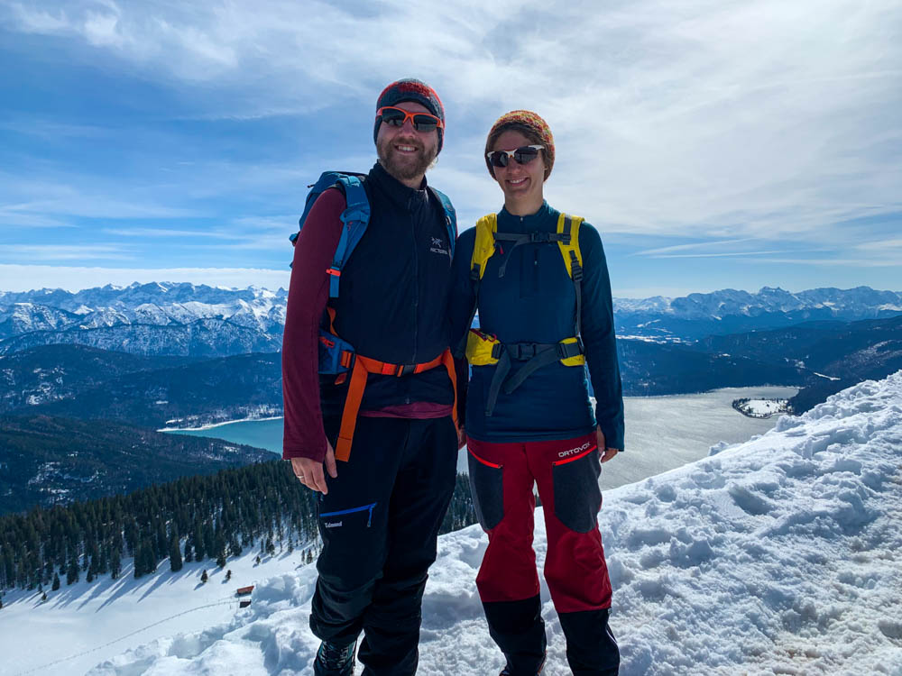 Melanie und Julian posieren am Gipfel des Jochberg. Hinter ihnen ist der Walchensee sowie eine schneebedeckte Berglandschaft zu sehen. Die Sonne in den Alpen lässt den Schnee strahlen. Reise Blog