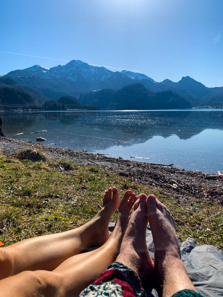 Melanie und Julian liegen am Kochelsee und lassen sich von der Sonne in den Alpen verwöhnen. Es sind ihre Füße im Vordergrund, der Kochelsee und dahinter dann eine Berglandschaft zu sehen.