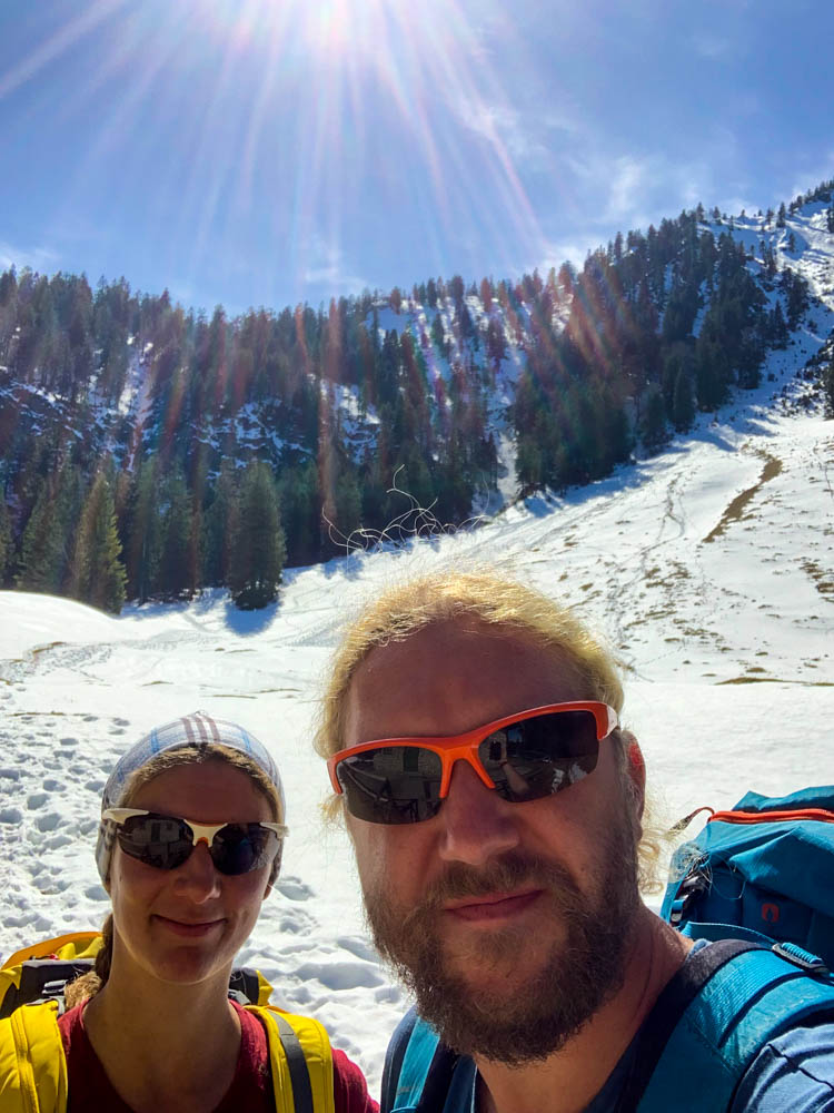 Selfie von Melanie und Julian von Orterer Hütte Richtung Rabenkopf. Hinter ihnen strahlt die Sonne in den Alpen und es ist ein Hang zu sehen, in dem bereits eine Lawine abgegangen ist.