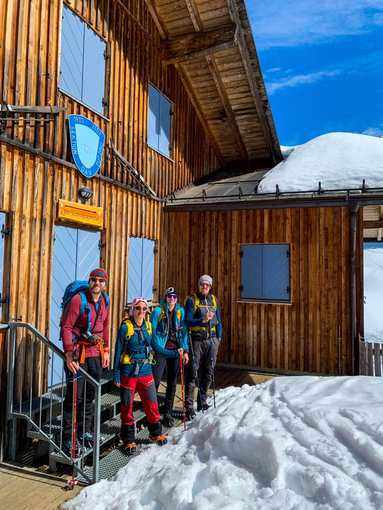 Melanie, Julian und zwei Freunde stehen vor der Tutzinger Hütte. Vor der Hütte liegt Schnee und der Himmel ist blau.