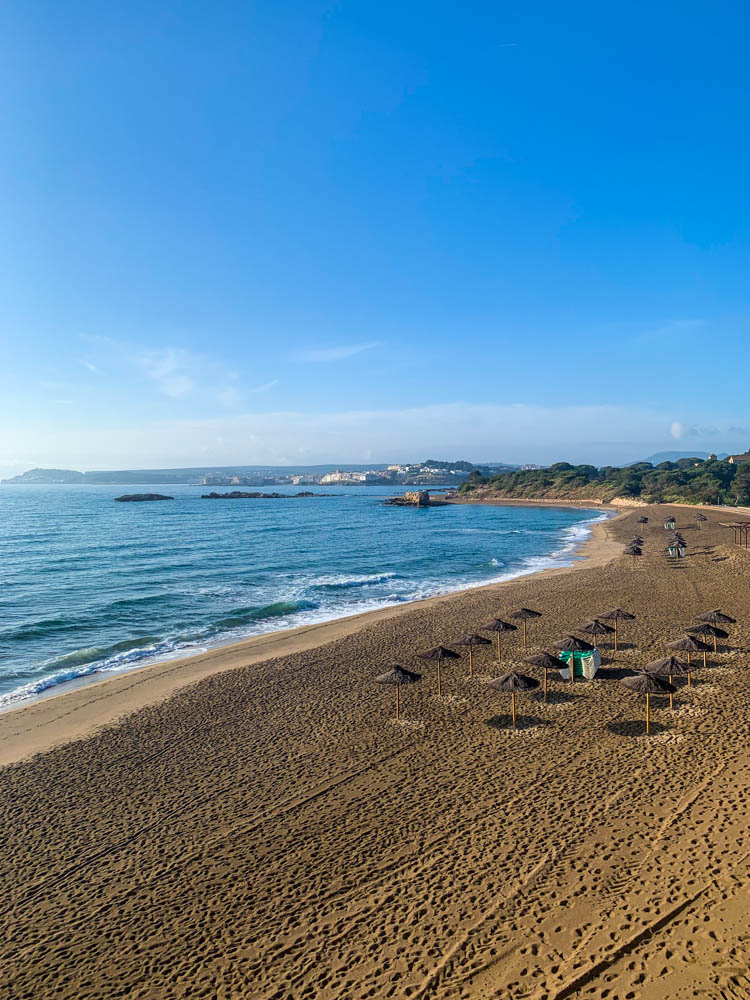 Ein Strand der Costa Brava am Mittelmeer. Das Bild ist von einem Plateau aufgenommen. im Sand sind viele Fußspuren zu sehen, es gibt ein paar Palmblättersonnenschirme und links im Bild rollen kleine Wellen des Mittelmeers an den Strand. Der Himmel strahl im hellsten blau.