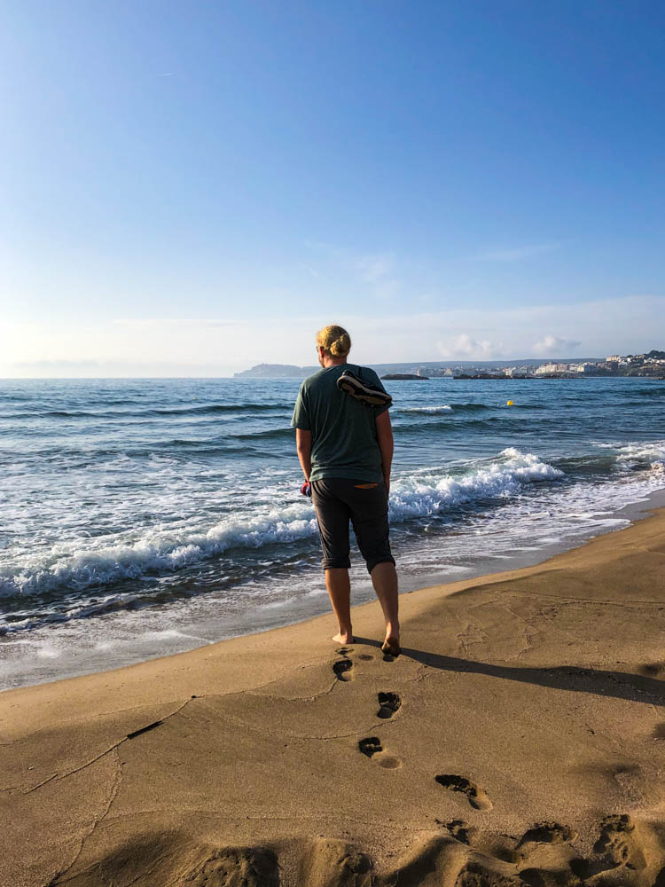 Julian läuft durch den weichen Sand an einem Strand der Costa Brava. Seine Fußabdrücke sind deutlich zu erkennen. Er steht ganz vorne am Wasser un die anrollende Welle wird ihm sicher nasse Füße bereiten.