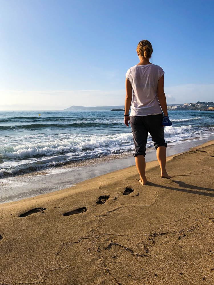 Melanie läuft durch den weichen Sand an einem Strand der Costa Brava. Ihre Fußabdrücke sind deutlich zu erkennen. Sie steht ganz vorne am Wasser un die anrollende Welle wird ihr sicher nasse Füße bereiten.