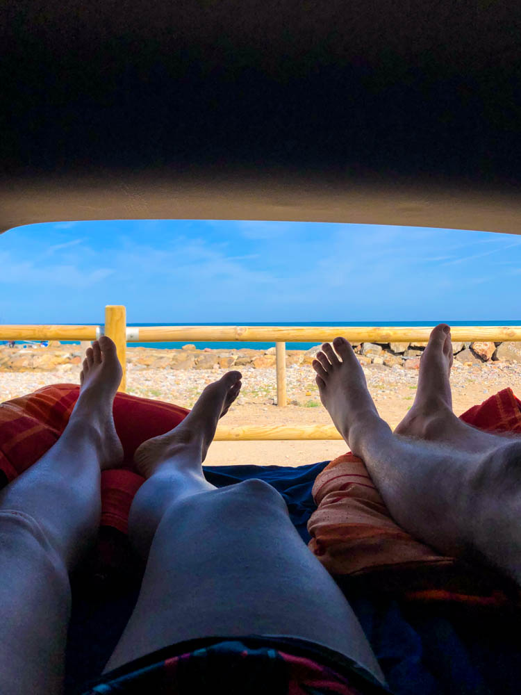 Blick aus unserem Mini-Camperauf unserem Weg auf die Kanaren. Wir liegen beide im Bett, die Heckklappe ist geöffnet und man sieht nur unsere Füße. Im Hintergrund ist das Mittelmeer und der Blaue Himmel zu sehen.