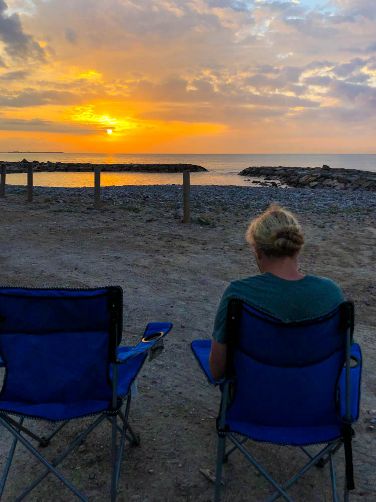 Unsere Campingstühle stehen beim Schlafplatz direkt am Mittelmeer auf dem Parkplatz. Im Hintergrund geht die Sonne unter und der Himmel leuchtet in einem schönen orange. Julian sitzt im rechten der beiden blauen Campingstühle und schaut auf das orange schimmernde Mittelmeer. Mit dem Mini-Camper auf die Kanaren