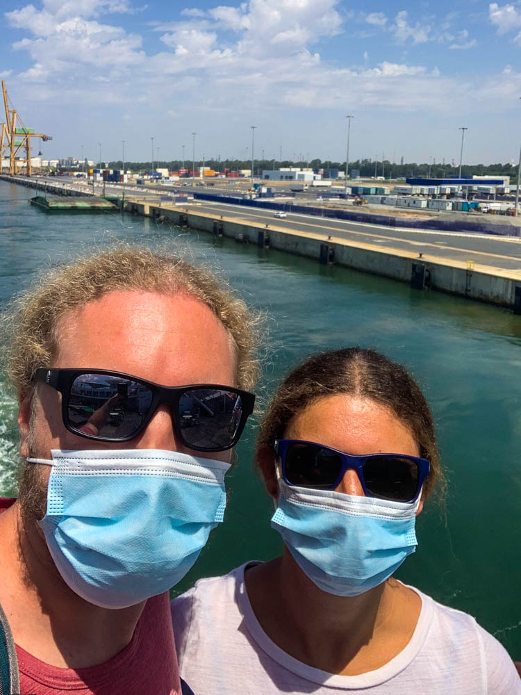 Selfie mit Sonnenbrille und Medizinische Maske von Melanie und Julian auf der Fähre. Im Hintergrund ist der Hafen von Huelva zu sehen. Das letzte Bild mit Festland sozusagen.