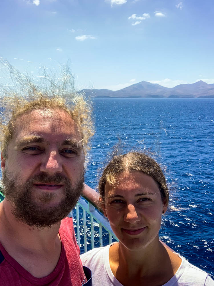 Selfie von Melanie und Julian auf der Fähre auf ihrem Weg mit dem Mini.Camper auf die Kanaren. Im Hintergrund ist das Festland der ersten Kanarischen Insel zu sehen. Sie haben ihr Ziel fast erreicht.