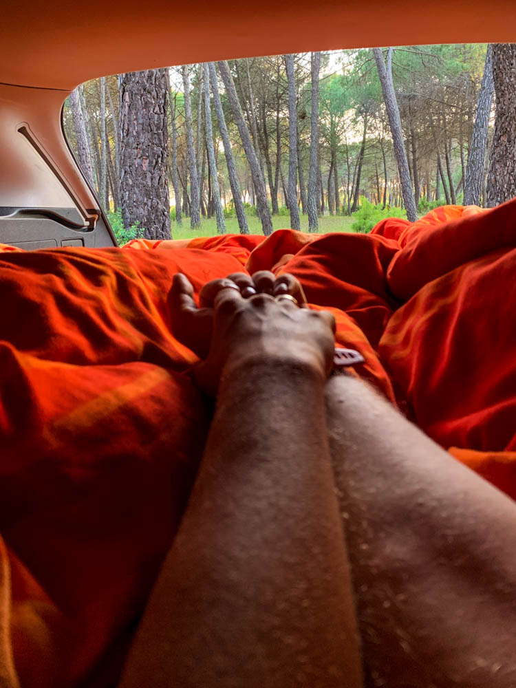 Julian und Melanie liegen in ihrem Mini-Camper auf dem Weg auf die Kanaren. Der Kofferraum ist geöffnet, sie halten Händchen und der Ausblick zeigt einen Pinienwald.