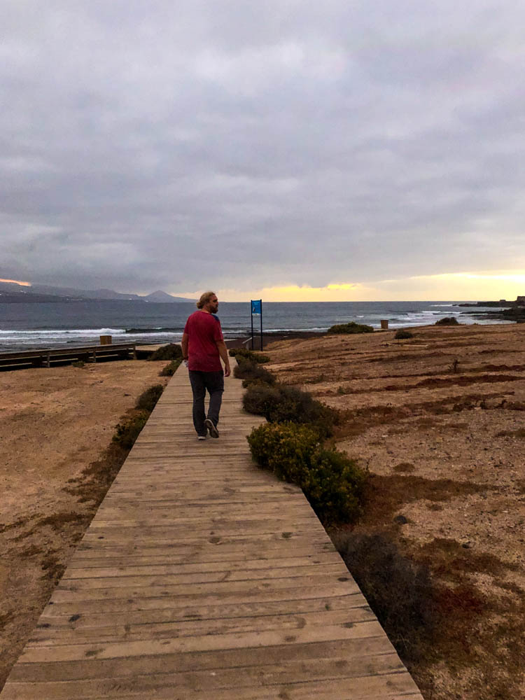 Julian läuft auf einem Steg Richtung Strand, welcher in der Nähe von Las Palmas liegt. Der Himmel ist vom Sonnenuntergang orange verfärbt