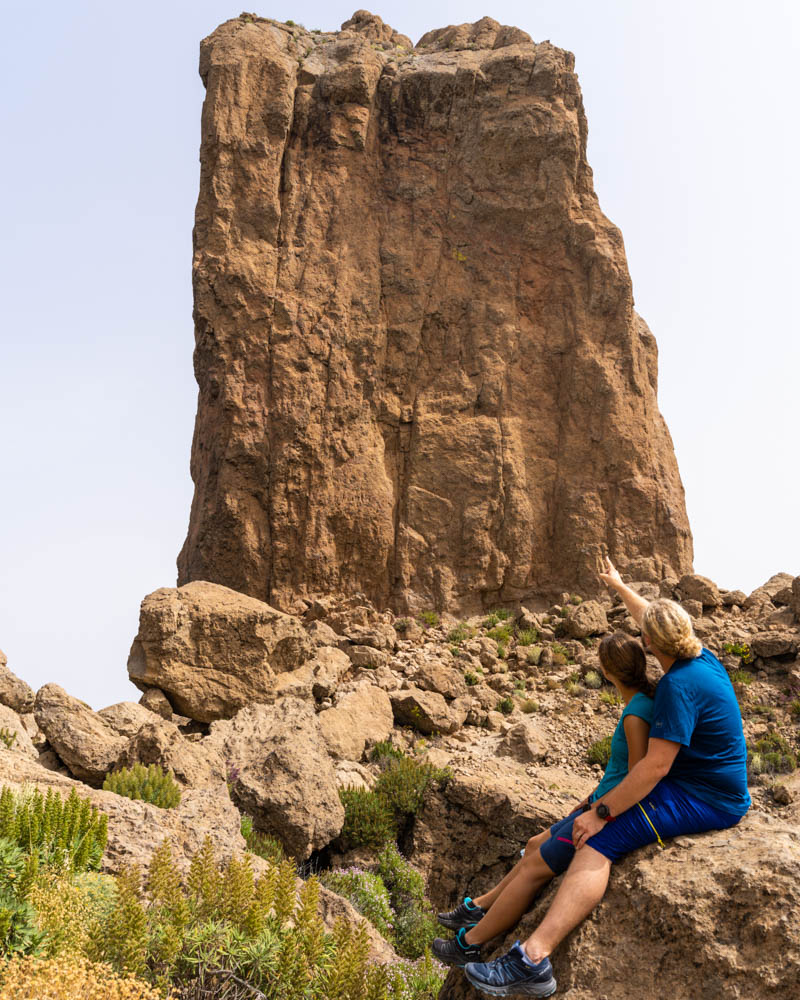 Melanie und Julian sitzen auf einem Stein vor Roque Nublo Gran Canaria. Julian zeigt mit seinem Finger auf den massiven Berg.