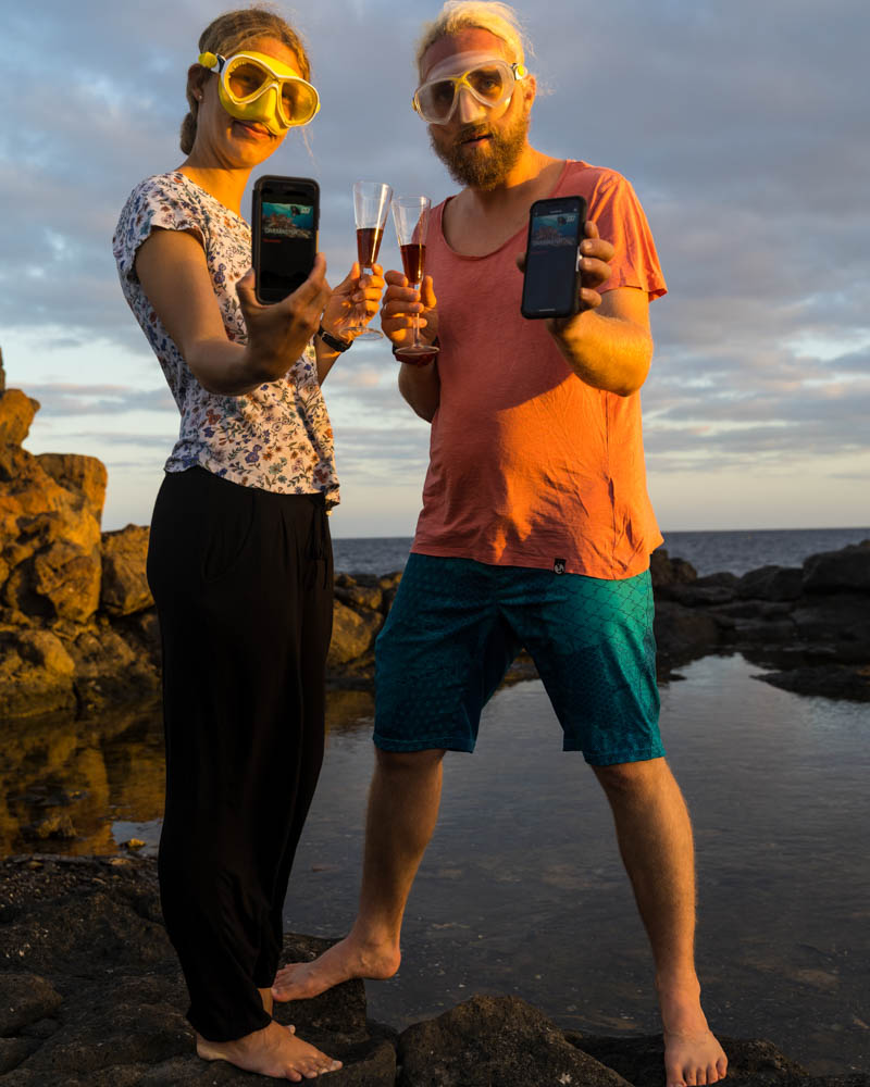 Melanie und Julian stehen beide am Atlantik, haben ihre Taucherbrillen auf und halten ihre Handys mit der Divemaster Zertifizierung ins Bild. Parallel stoßen sie mit zwei Sektgläsern auf das bestandene Divemaster Praktikum an.