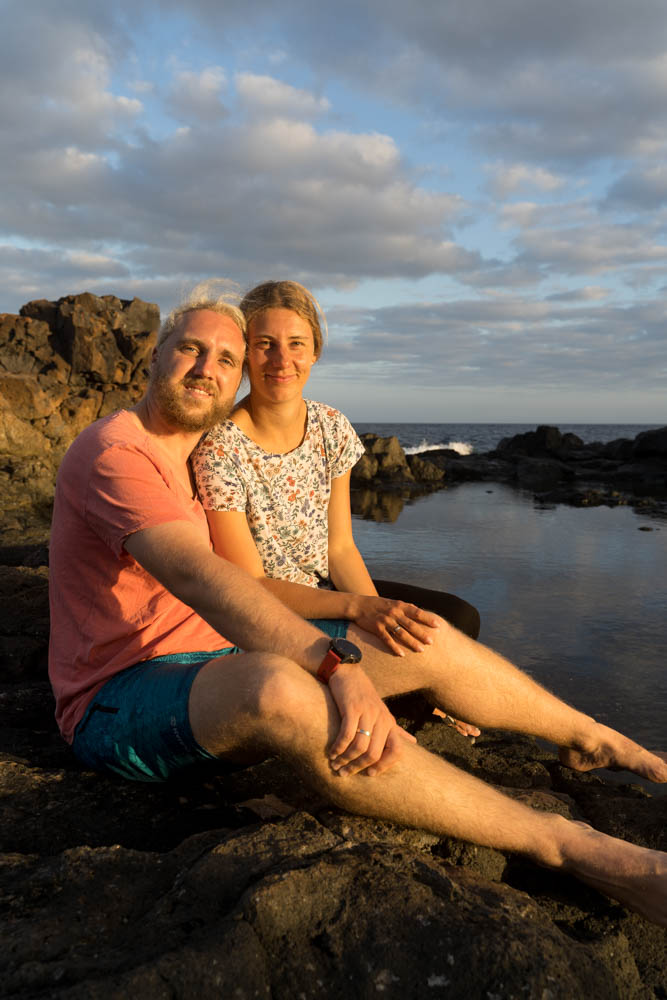 Melanie und Julian sitzen an der Atlantikküste und lächeln als schönes Paar in die Kamera.