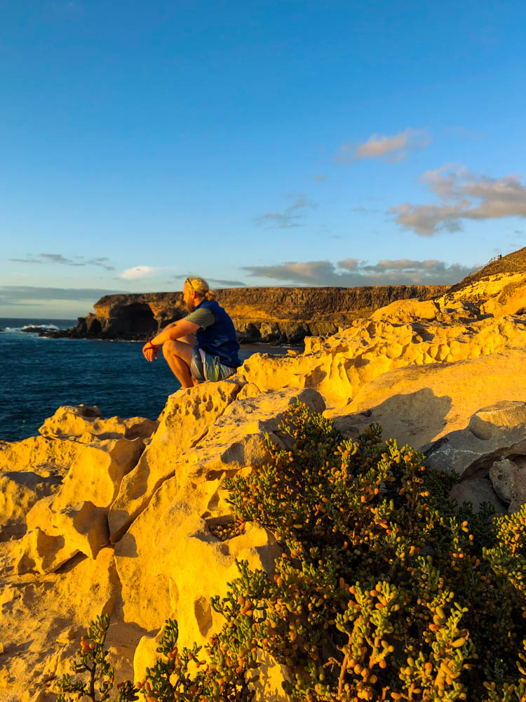Julian sitzt an den Klippen bei Ajuy und schaut auf den Atlantik.Der Gelbe Sandfelsen bietet einen starken Kontrast zu blauem Himmel und Atlantik.