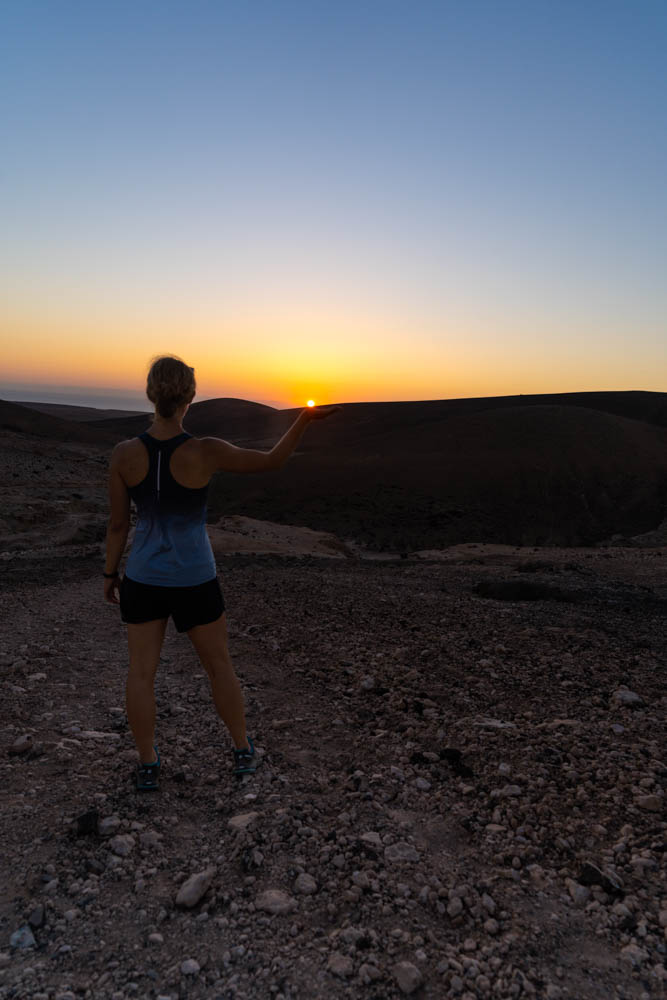 Melanie steht während dem Sonnenuntergang auf einem Hügel und streckt die Hand aus. Es sieht so aus als würde sie die untergehende rote Sonnenscheibe in ihrer Hand halten. Fuerteventura entdecken kann man zu jeder Tageszeit.