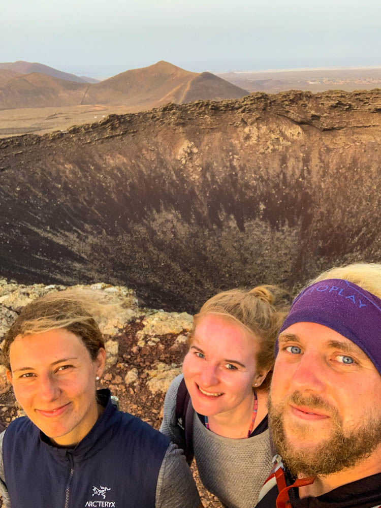 Lara, Melanie und Julian stehen auf dem Vulkan Calderon Hondo. Im Hintergrund ist der große Krater zu sehen. Am Himmel ist der viele Saharastaub durch Calima zu sehen. Auch Calima kann man während eines Fuerteventura Urlaubs entdecken.