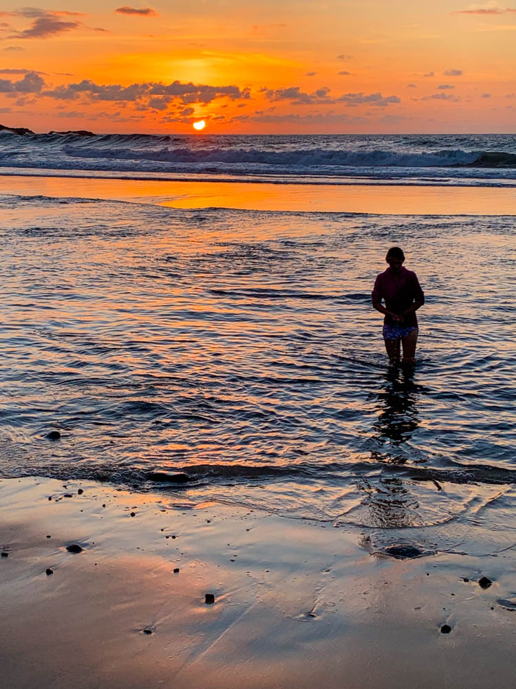 Sonnenuntergang Los Molinos. Melanie läuft durchs Wasser und hinter ihr geht die Sonne unter. Der Himmel und das Meer sind orange-rot.