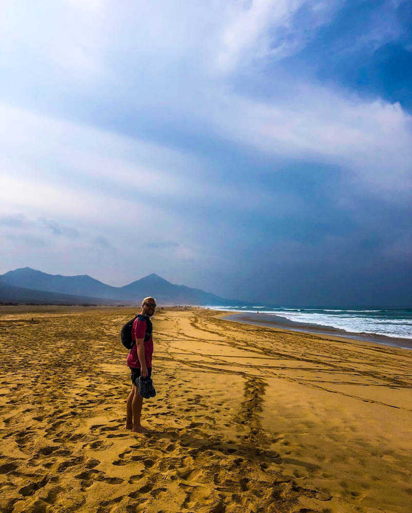 Julian bei Wanderung nach Cofete. Julian steht barfüßig am kilometerlangen Strand, der Himmel ist bewölkt. Wandernd schöne Ecken auf Fuerteventura entdecken.