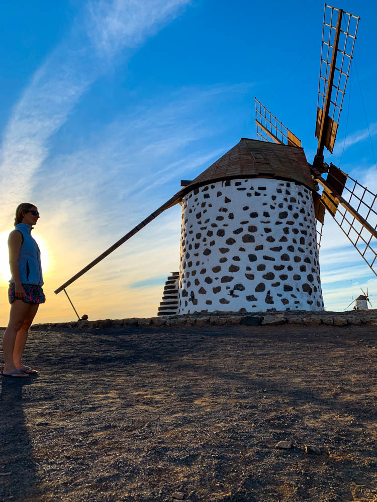 Melanie steht vor einer Windmühle auf Fuerteventura. Im Hintergrund ist noch eine weitere Windmühle zu sehen. Der Himmel ist intensiv blau und leicht orange verfärbt.