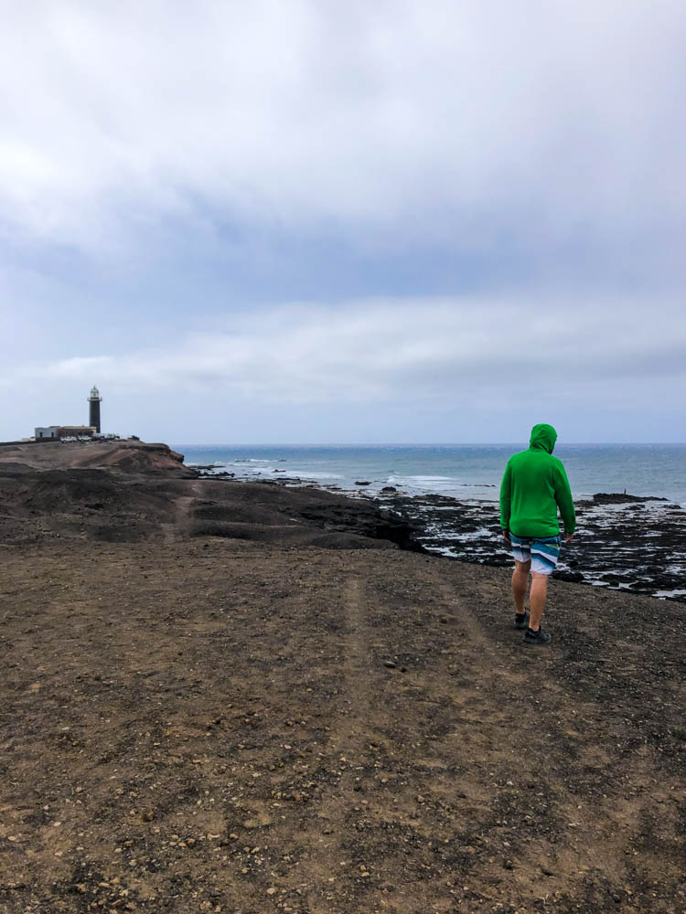 Julian geht an der südlichsten Stelle auf Fuerteventura spazieren. Im Hintergrund ist der Leuchtturm Punta Jandia zu sehen. Der Himmel ist bewölkt.