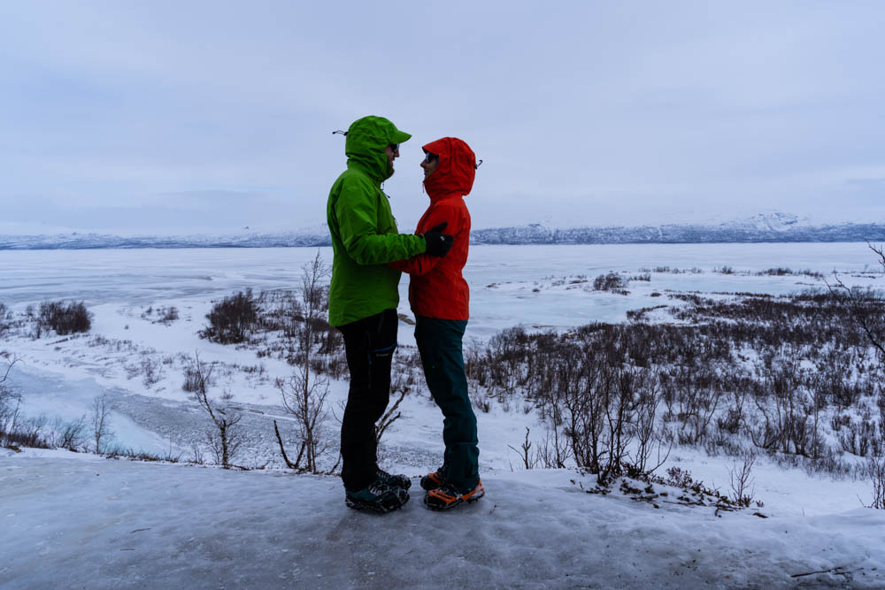 Melanie und Julian stehen sich auf Eis und Schnee gegenüber und blicken sich tief in die Augen. Hinter ihnen ist der Torneträsk-See zu sehen. Abisko Nationalpark Schweden