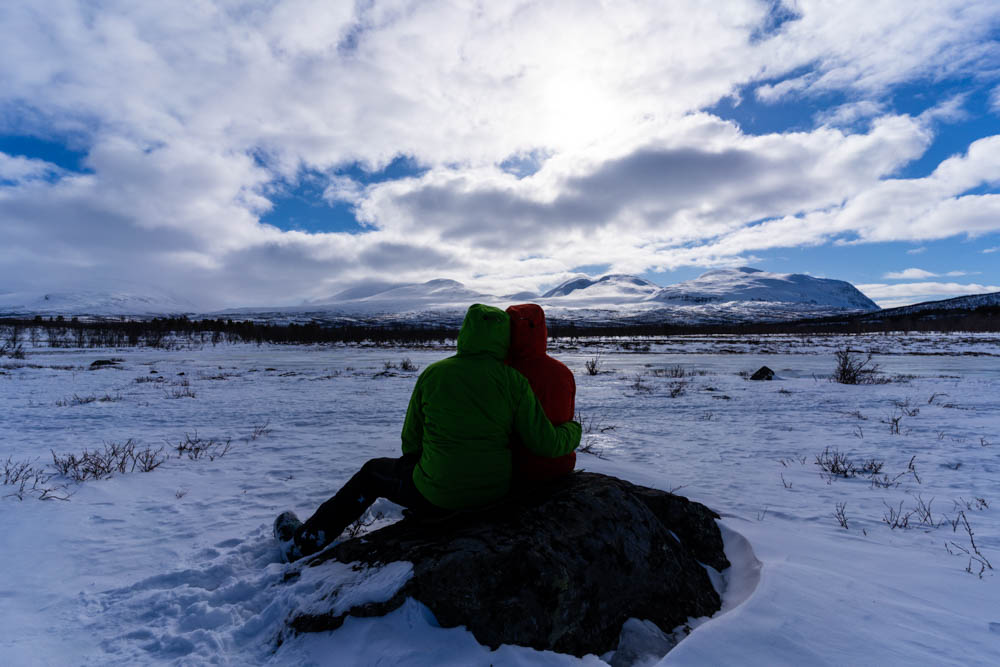 Melanie und Julian sitzen auf einem Stein und blicken in die Ferne. Schöne Schneelandschaft mit Bergen und einem See im Abisko Nationalpark Schweden