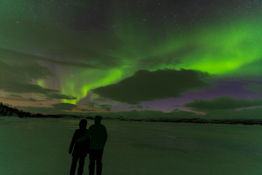 Melanie und Julian stehen auf dem See Torneträsk, über ihnen tanzen Nordlichter. Abisko Nationalpark Schweden