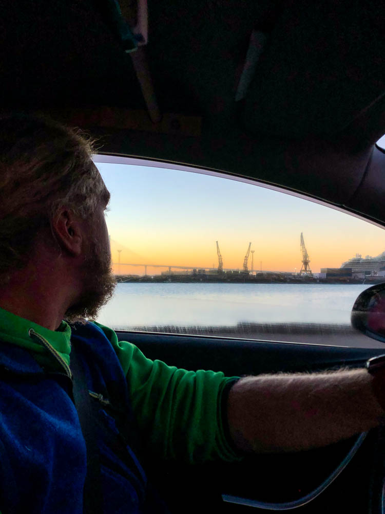 Julian sitzt am Steuer von Mini-Camper Astrarix und fährt gerade von der Fähre. Er blickt nach draußen, wo gerade die Sonne aufgeht und der Hafen zu sehen ist.