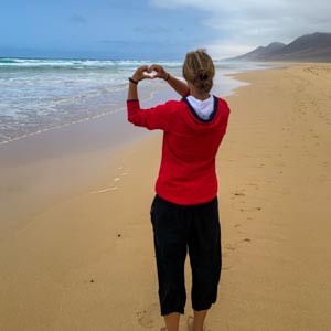 Fuerteventura entdecken - Melanie formt mit ihren Händen ein Herz, sie steht am ewig langen Sandstrand in Cofete und blickt auf das Meer.