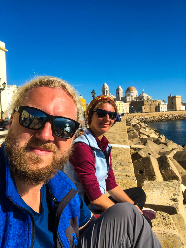 Melanie und Julian sitzen auf einer Mauer direkt am Atlantik. Im Hintergrund ist u.a. die Kathedrale sowie der Dom von Cadiz zu sehen. Der Himmel ist strahlend blau.