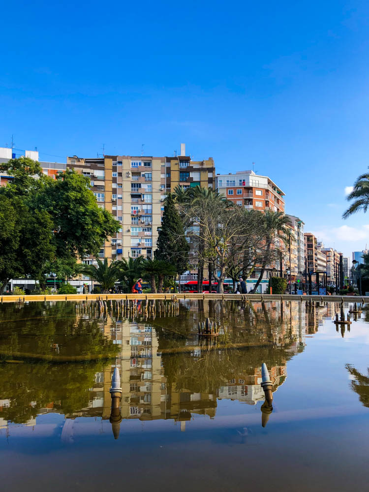 Häuser der Stadt Murcia spiegeln sich in einem großen Teich wider. Der Himmel ist kräftig blau.