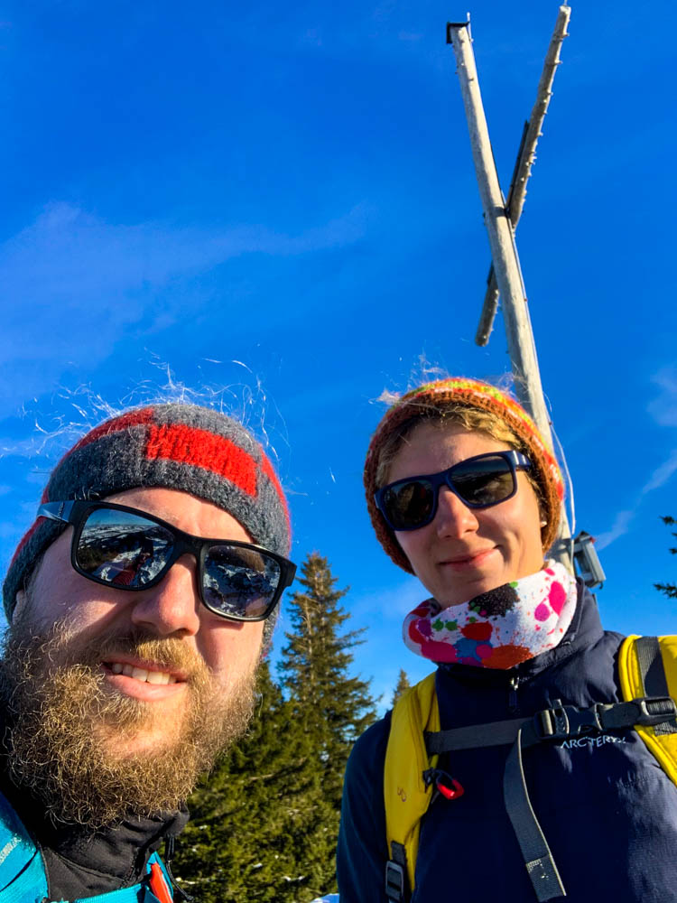 Melanie und Julian posieren mit dem Gipfelkreuz der Alpspitze bei Nesselwang. Der Himmel ist kräftig blau. Winter in den Alpen - Gipfelerfolg
