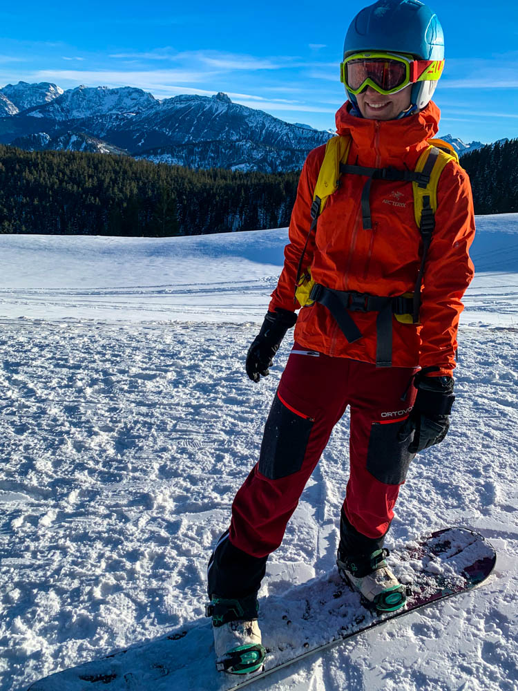 Melanie grinst in die Kamera und steht gerade auf ihrem als Board zusammengebautem Splitboard. Sie ist von Schnee umgeben, der Himmel ist kräftig blau und im Hintergrund sind die Berge. Durch ihr Outfit leuchtet Melanie schön bunt heraus und bietet einen schönen Kontrast.