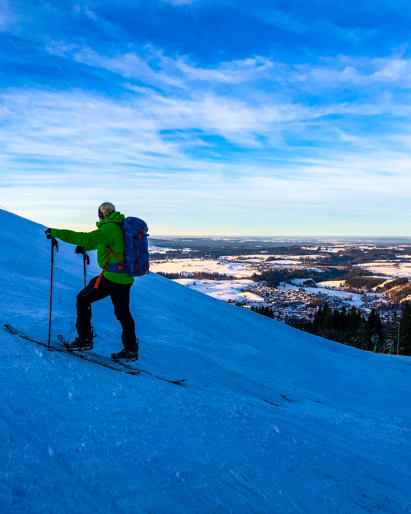 Julian im Aufstieg mit seinem Splitboard. Er schaut gerade auf Nesselwang und die schöne Schneelandschaft hinab. Der Himmel leuchtet kräftig blau.