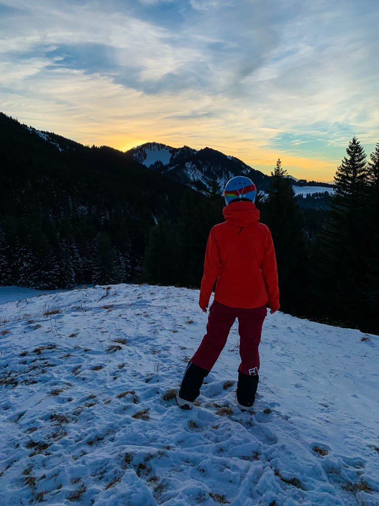 Melanie kurz vor der Abfahrt mit ihrem Splitboard bei Nesselwang. Sie schaut auf die gegenüberliegenden Berge, wo gerade die Sonne untergeht. Winter in den Alpen - Sonnenuntergang