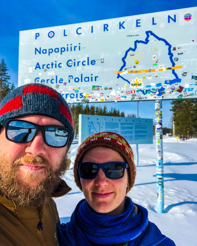 Selfie von Melanie und Julian vor Polarkreis Schild in Schweden. Der Himmel ist strahlend blau, der Boden ist mit Schnee bedeckt. Fahrt in den hohen Norden über Schweden