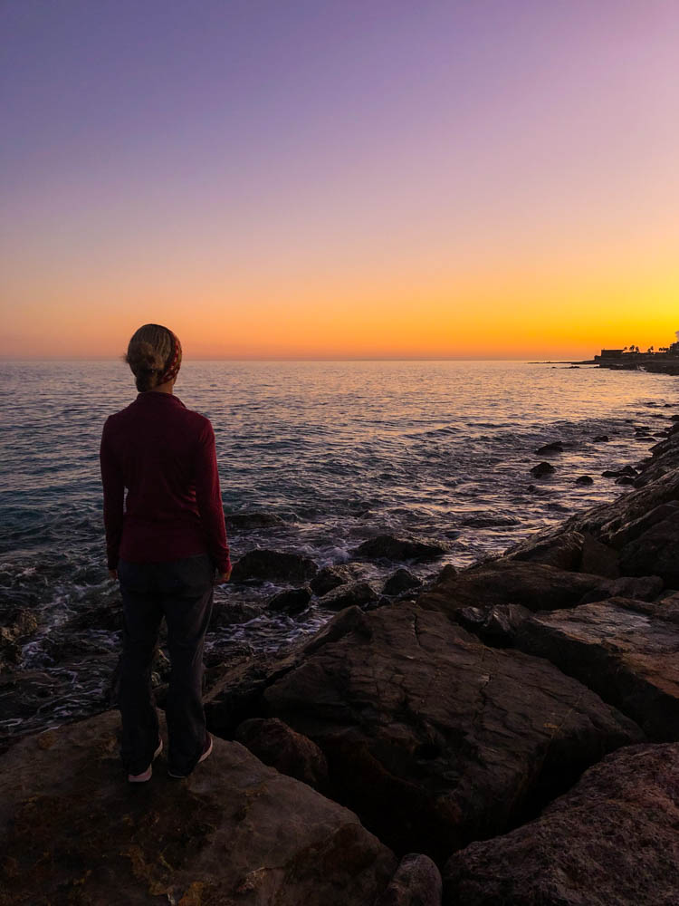Melanie steht auf einer kleinen Steinmauer und blickt auf das Mittelmeer. Die Sonne ist gerade untergegangen und der Himmel ist orange verfärbt.
