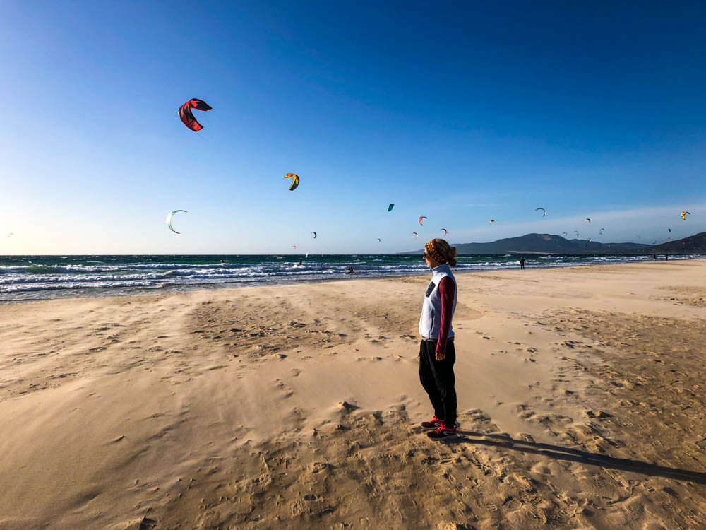 Melanie steht am Strand bei Tarifa und blickt auf den Atlantik. Am strahlend blauen Himmel sind etliche bunte Schirme von Kitesurfern zu sehen. Rückfahrt über Spanien - Tarifa