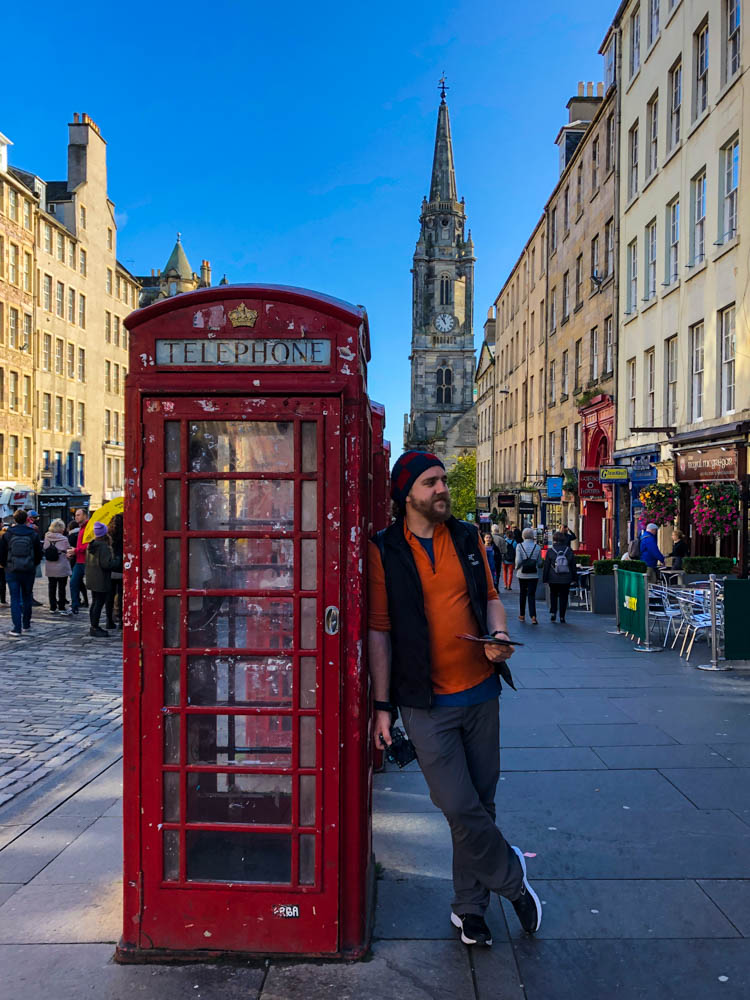 Julian lehnt in Edinburgh an einer roten Telefonzelle. Er steht in der Innenstadt, es ist eine Kirche sowie einige Gebäude zu sehen. Der Himmel ist strahlend blau.