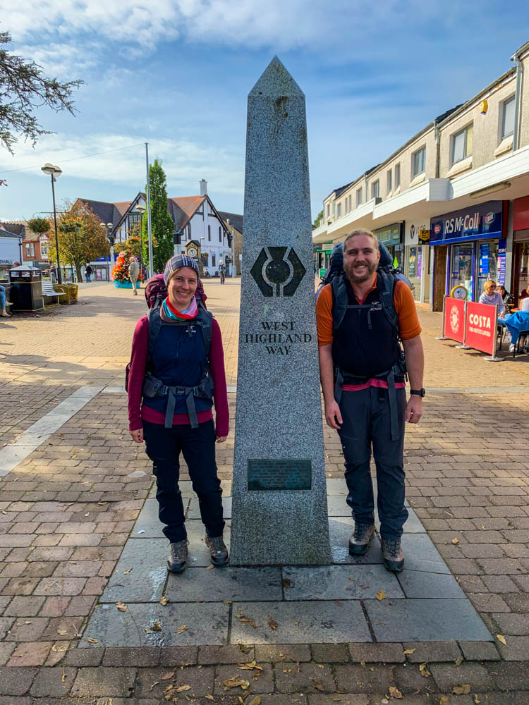 Melanie und Julian stehen neben einer Säule in Milngavie. Start des Trekking vom West Highland Way in Schottland.