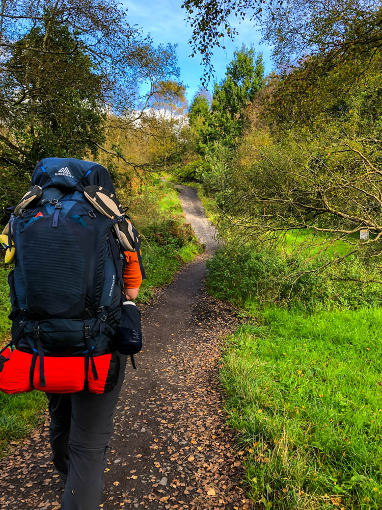 Julian wandert mit seinem vollgepackten Rucksack auf dem West Highland Way in Schottland. Er läuft durch ein Waldstück, alles ist schön grün.