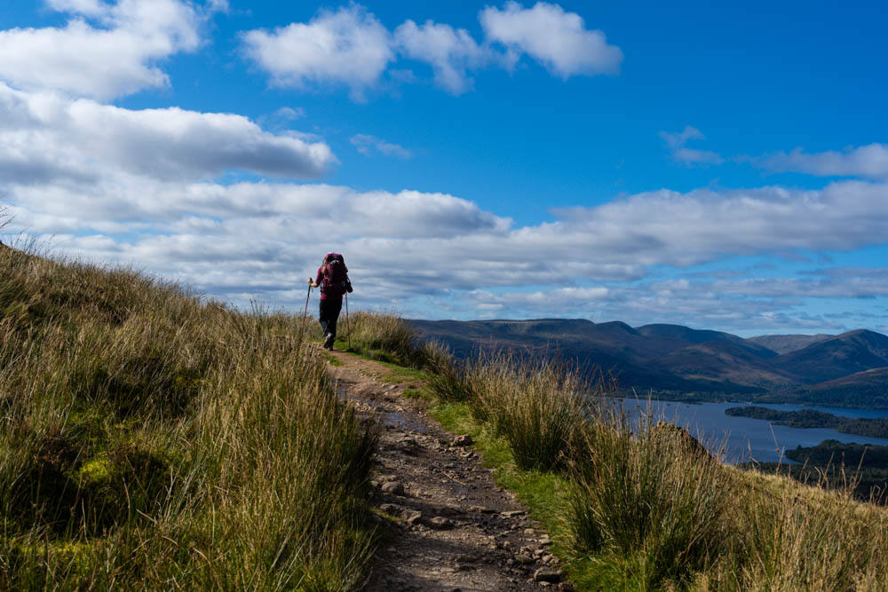 Melanie auf dem West Highland Way. Die Aussicht geht über Hügellandschaften und See hinaus. Der Himmel ist leicht bewölkt